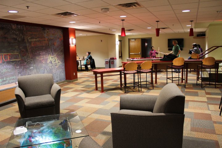 A lounge area with armchairs, a glass coffee table, and a large chalkboard wall filled with colorful writing. In the background, people play pool, and one person is seated at a table working on a laptop. The room is brightly lit with overhead lights and red pendant lamps.